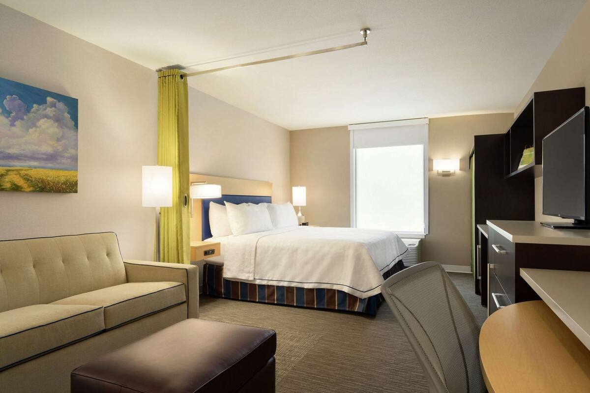 A hotel room with a bed, a sofa, a desk, a television, and a painting on the wall.