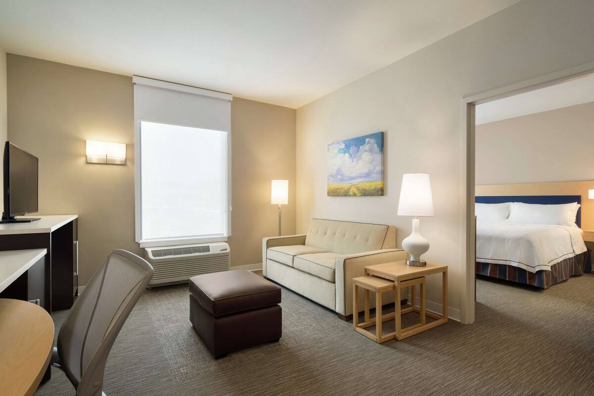 A hotel room with a desk, chair, sofa, ottoman, bedside table, lamp, and a bed.