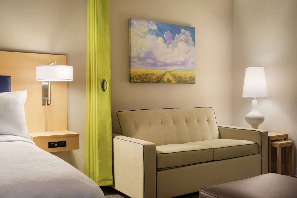 A hotel room with a bed, a sofa, a bedside table with a lamp, a painting on the wall, and a green curtain.