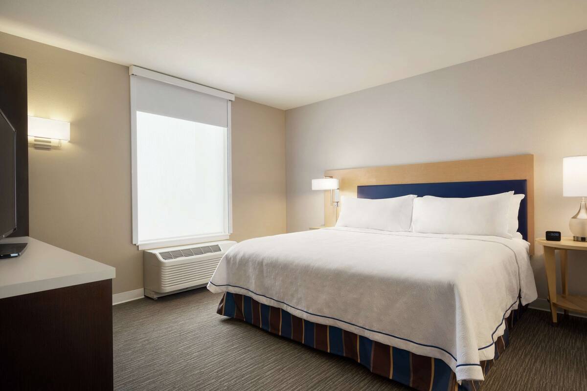 A hotel room with a large bed, a desk, a chair, a TV, a window with blinds, and a bedside lamp.