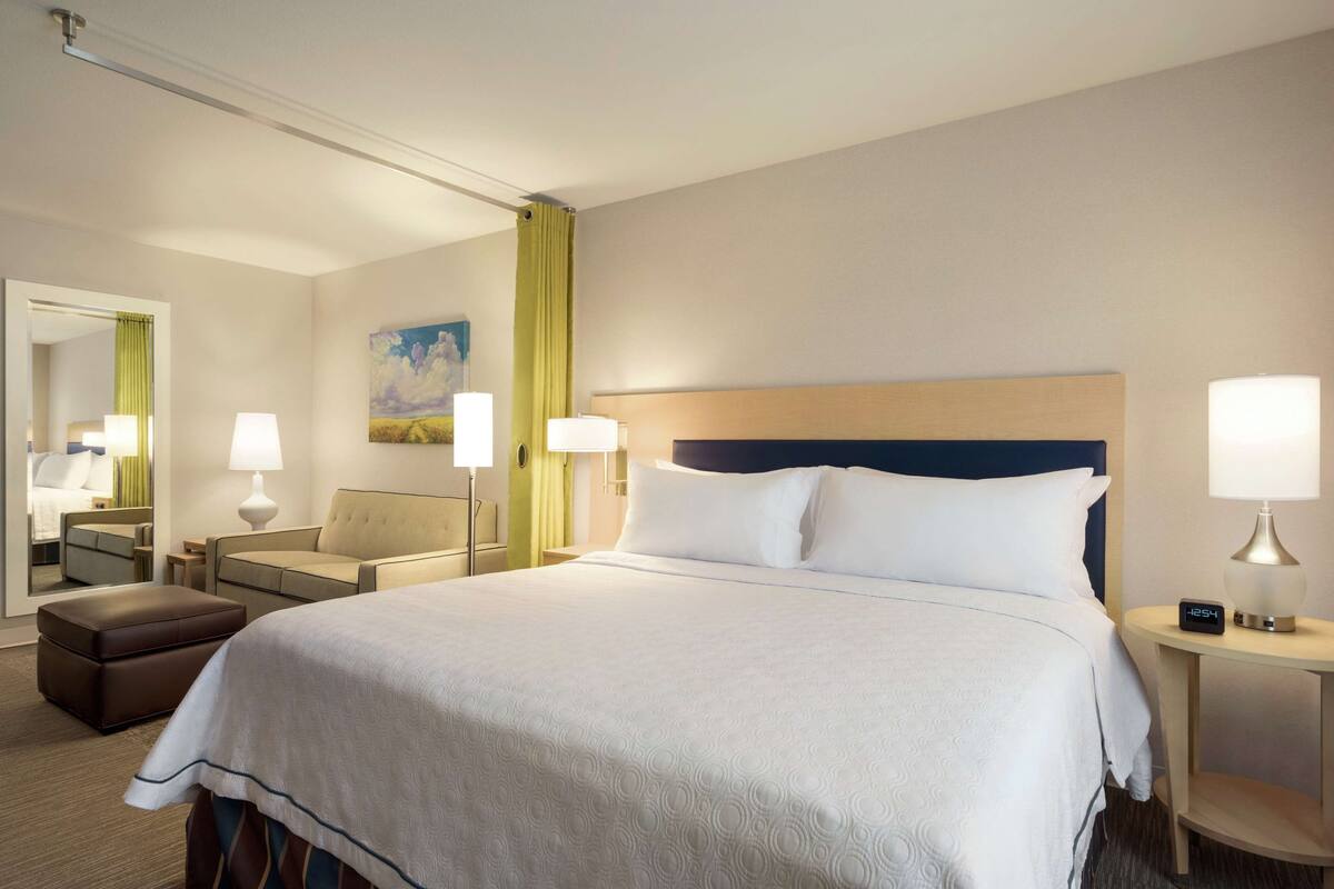 A hotel room with a large bed, two bedside tables with lamps, a nightstand, a chair, a mirror, and a painting on the wall.