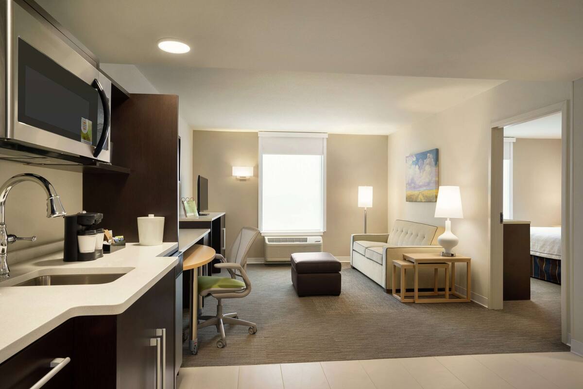 A modern hotel room with a kitchenette, a sofa, a small table with a lamp, and a window with a view.