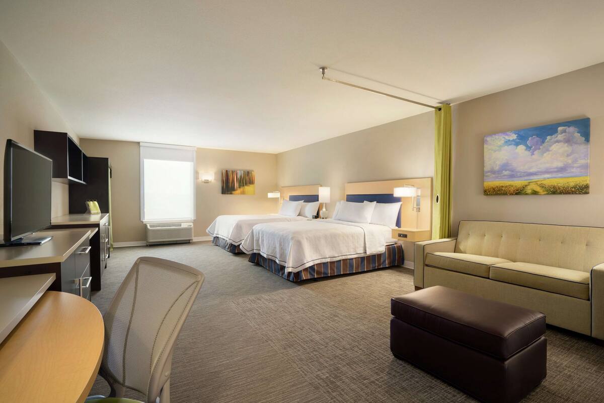 A hotel room with two beds, a sofa, a desk with a TV, and a painting on the wall.