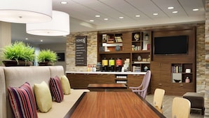A modern interior with a wooden dining table, cushioned seating, and a kitchen area with built-in shelves and a flat-screen TV. - Home2 Suites by Hilton Leavenworth Downtown (Leavenworth)