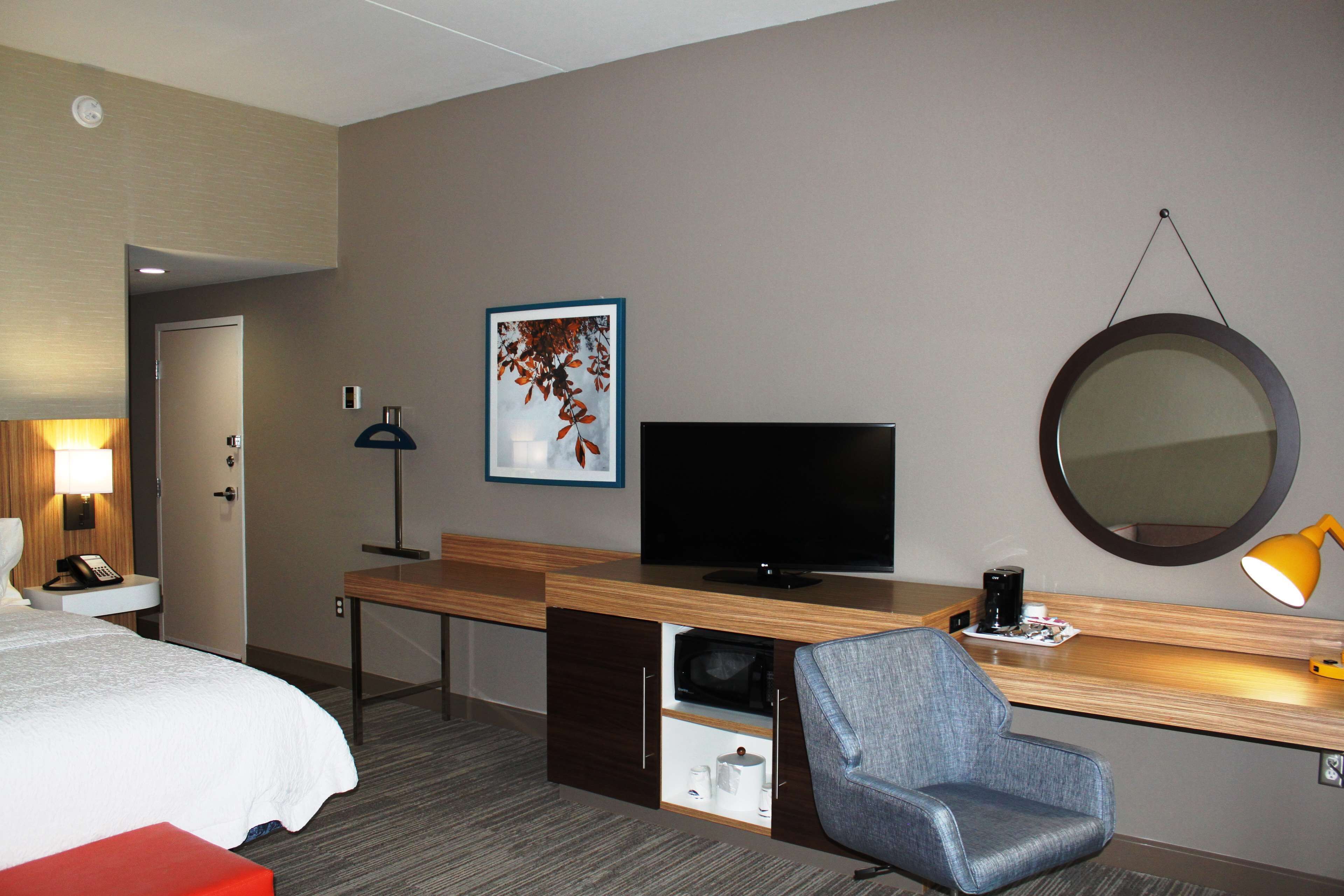 A hotel room with a bed, a desk with a TV, a chair, a desk lamp, a microwave, and a framed picture on the wall.