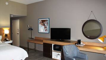 A hotel room with a bed, a desk with a TV, a chair, a desk lamp, a microwave, and a framed picture on the wall.