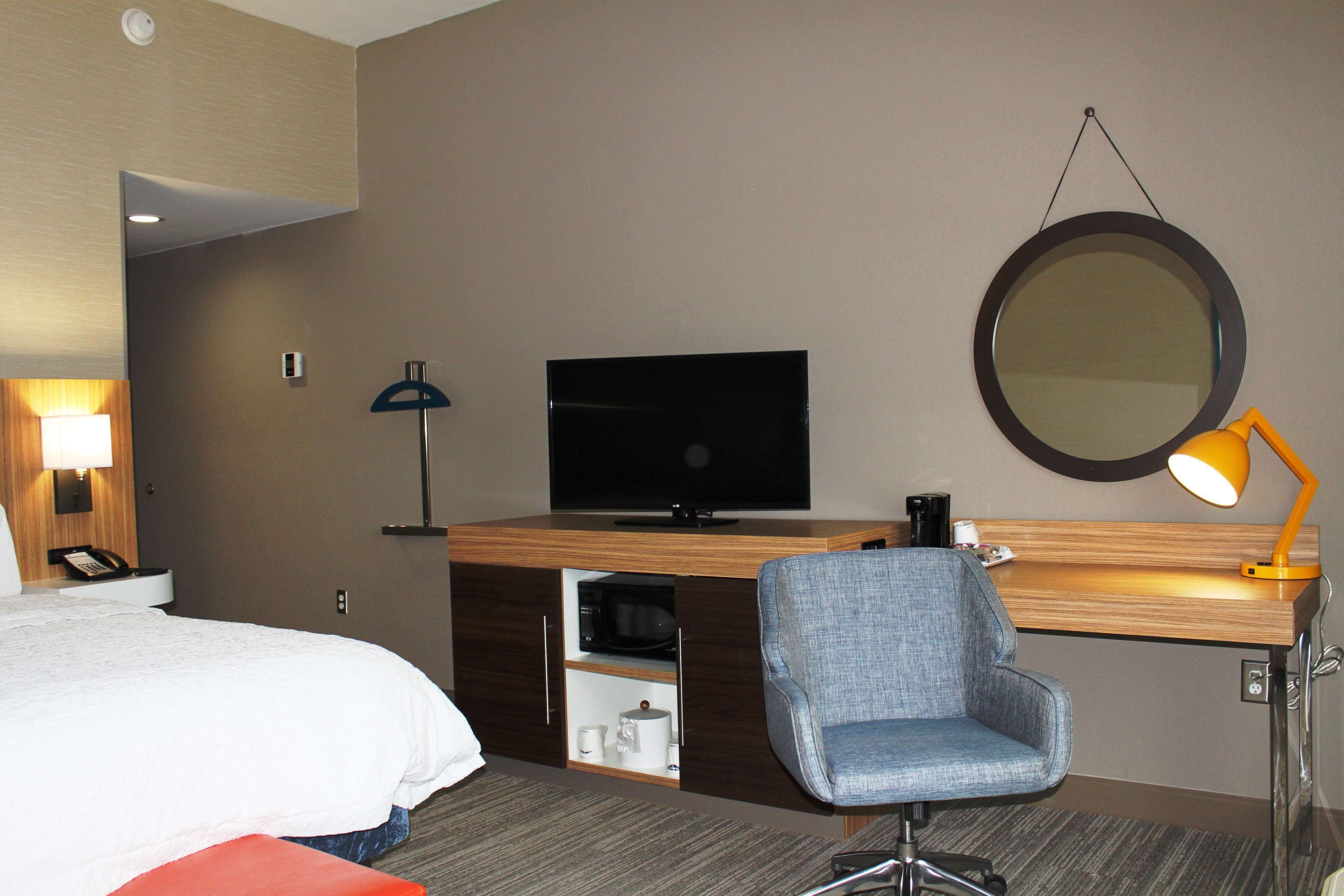 Quarto de hotel com cama, escrivaninha com televisão de tela plana, cadeira, abajur, espelho e máquina de café.