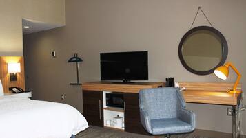 Quarto de hotel com cama, escrivaninha com televisão de tela plana, cadeira, abajur, espelho e máquina de café.