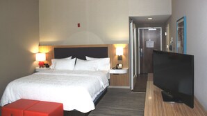 A hotel room with a bed, bedside lamps, a TV, and a small red ottoman.