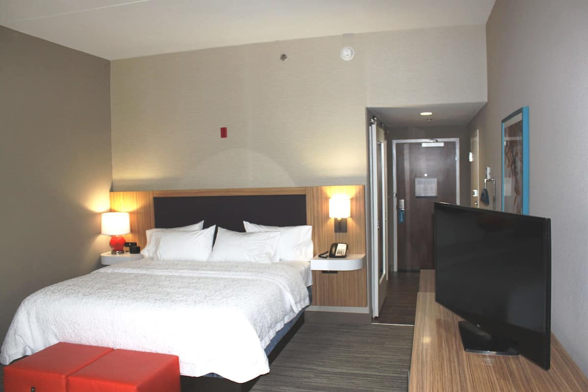 A hotel room with a bed, bedside lamps, a TV, and a small red ottoman.