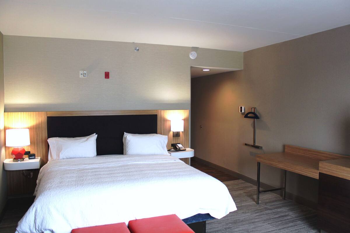 A hotel room with a large bed, bedside tables with lamps, a desk, and a bench.