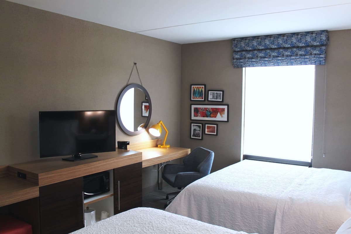 A hotel room with two beds, a desk with a television, a mirror, a lamp, and framed pictures on the wall.