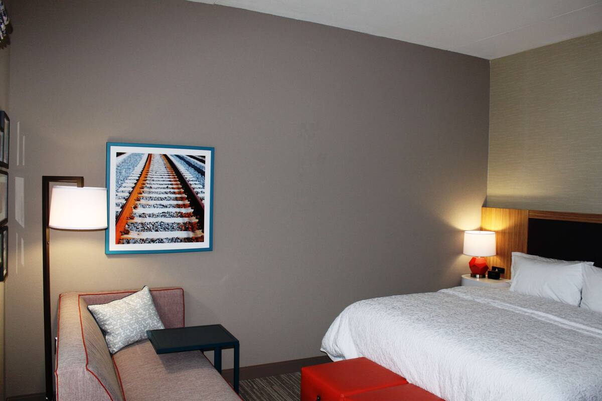 A hotel room with a bed, a sofa, a small table, a nightstand with a lamp, and a framed picture on the wall.