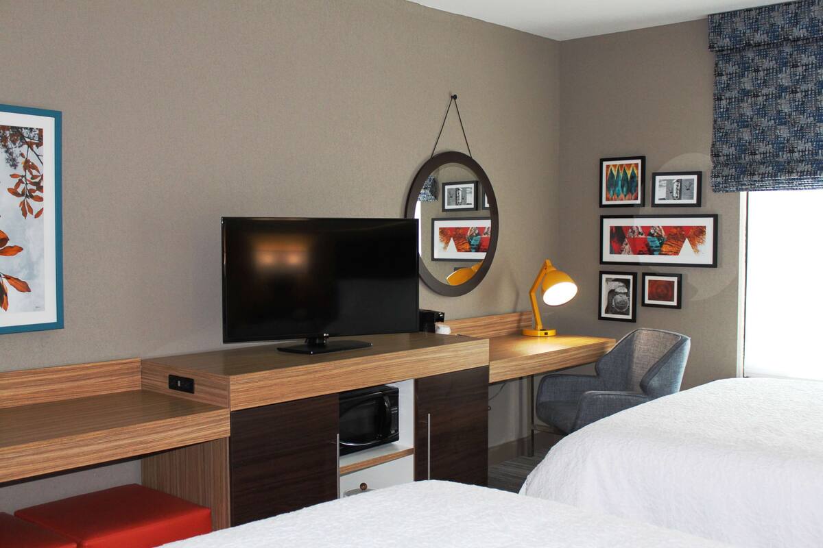 A hotel room with a wooden desk, a television, a mirror, a lamp, and a bed.