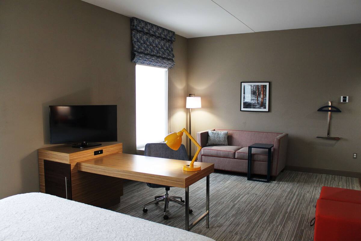 A hotel room with a wooden desk, a television, a sofa, a red chair, a lamp, and a framed picture on the wall.