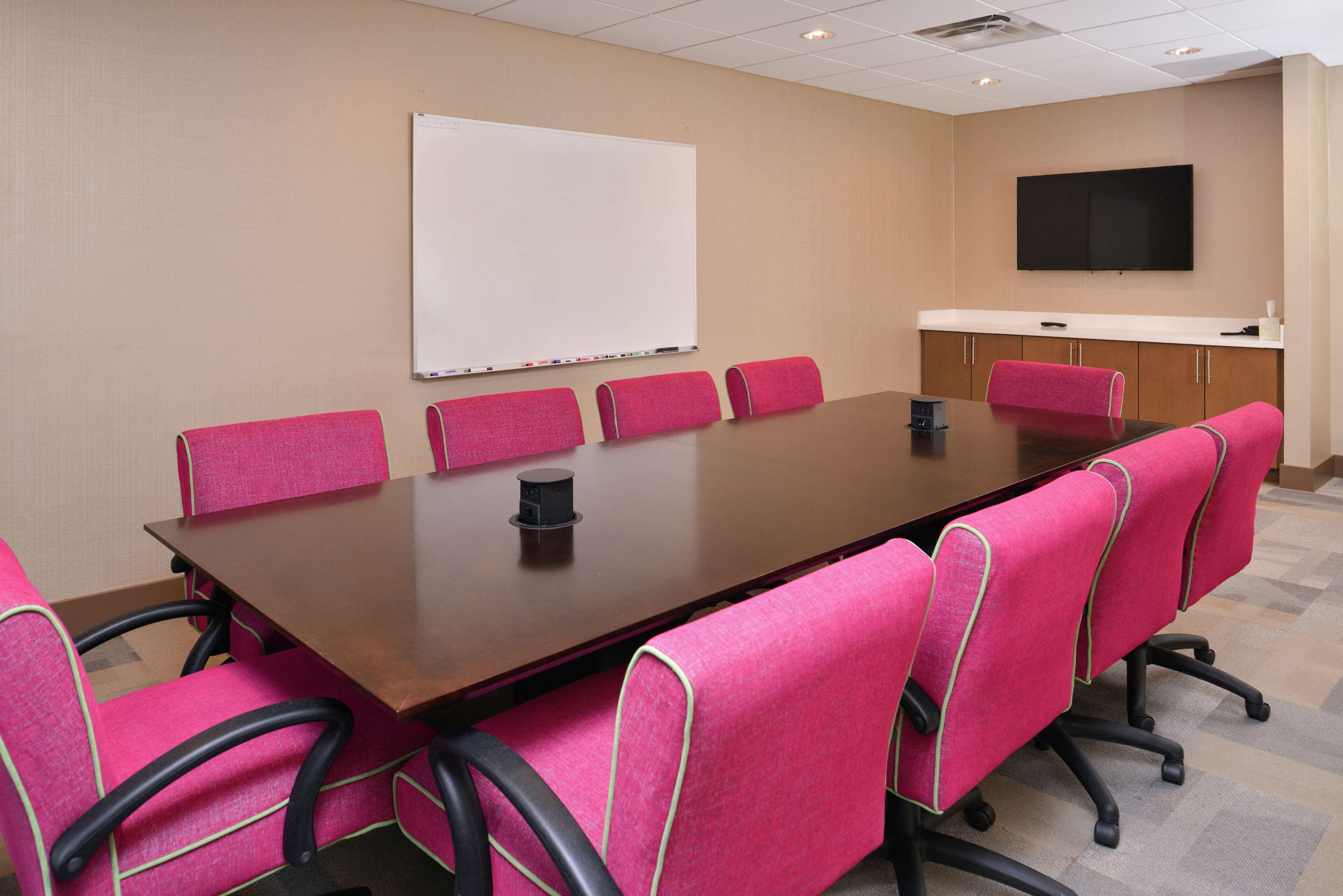 A conference room with a large table, six pink chairs, a whiteboard, and a flat-screen TV on the wall.
