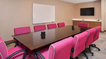 A conference room with a large table, six pink chairs, a whiteboard, and a flat-screen TV on the wall.