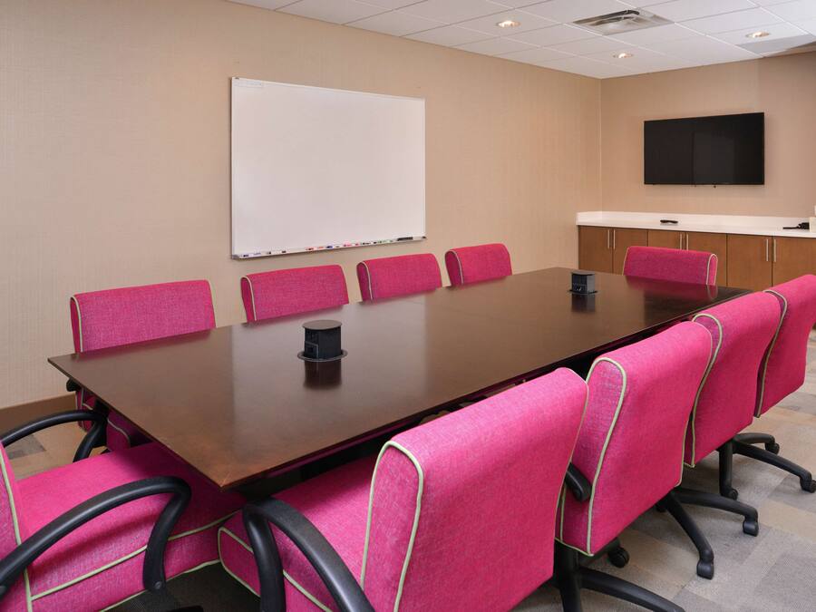 A conference room with a large table, six pink chairs, a whiteboard, and a flat-screen TV on the wall.