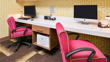 A hotel room with a desk, two chairs, two monitors, a keyboard, a mouse, a printer, and a lamp.