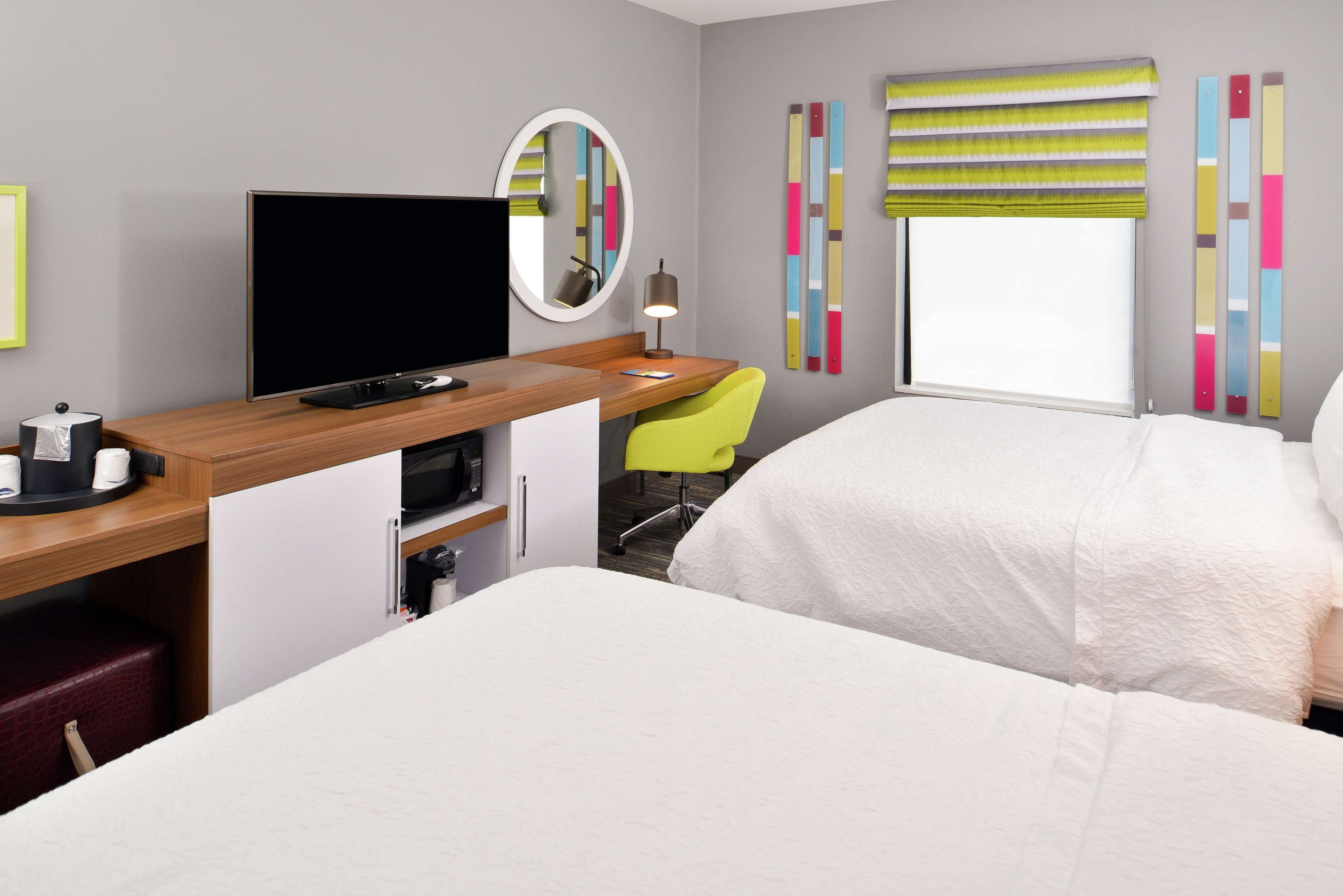 Quarto de hotel com cama, escrivaninha com computador, cadeira, televisão e janela com persianas.