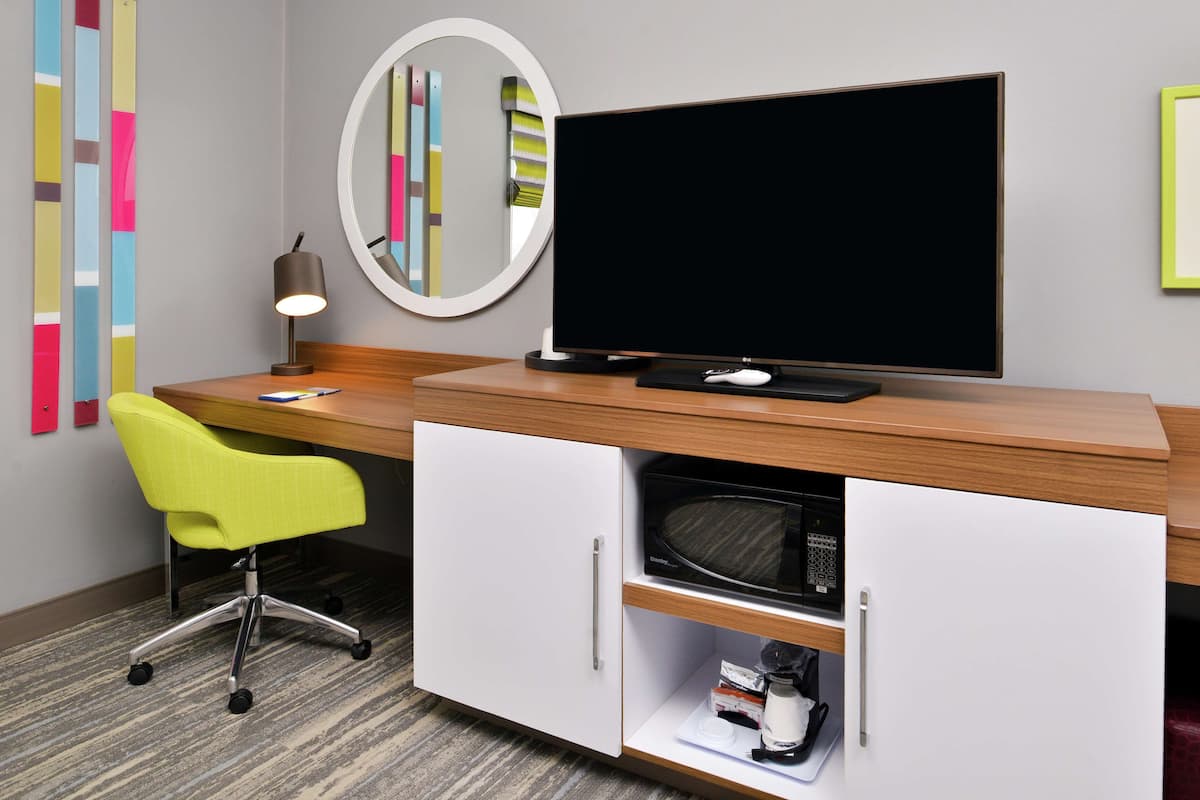 A hotel room with a wooden desk, a flat-screen TV, a yellow office chair, a mirror, and a microwave.