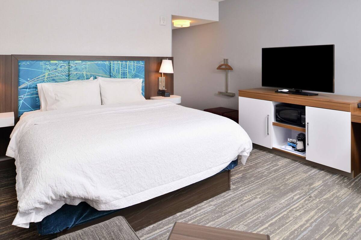 Habitación de hotel con cama, televisión, escritorio con microondas y lámpara de pared.