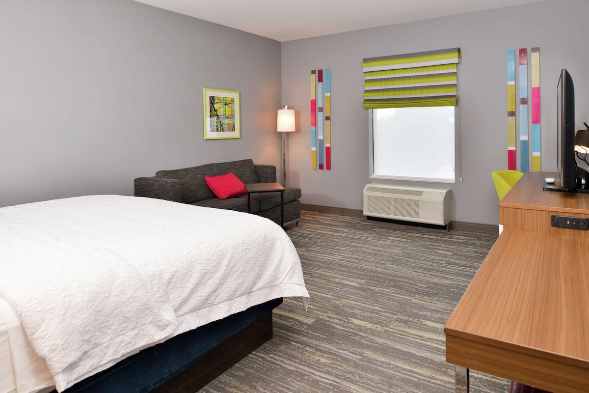 A hotel room with a bed, a desk, a chair, a lamp, a window with blinds, and a radiator.