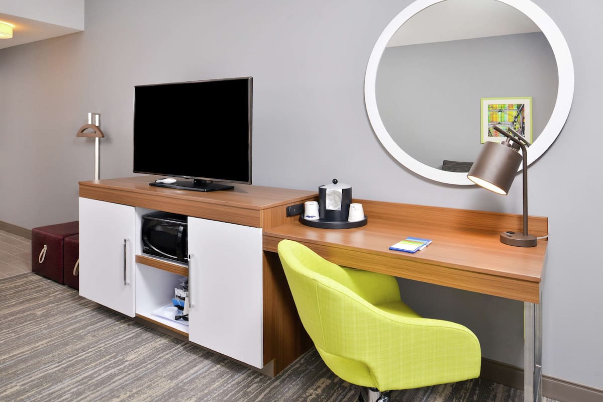A hotel room with a wooden desk, a television, a mirror, a green chair, and a red suitcase.