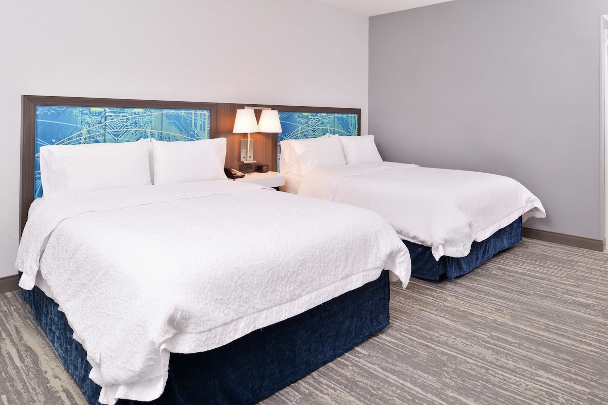 A hotel room with two beds, a headboard with a cityscape design, a nightstand with a lamp, and a patterned carpet.