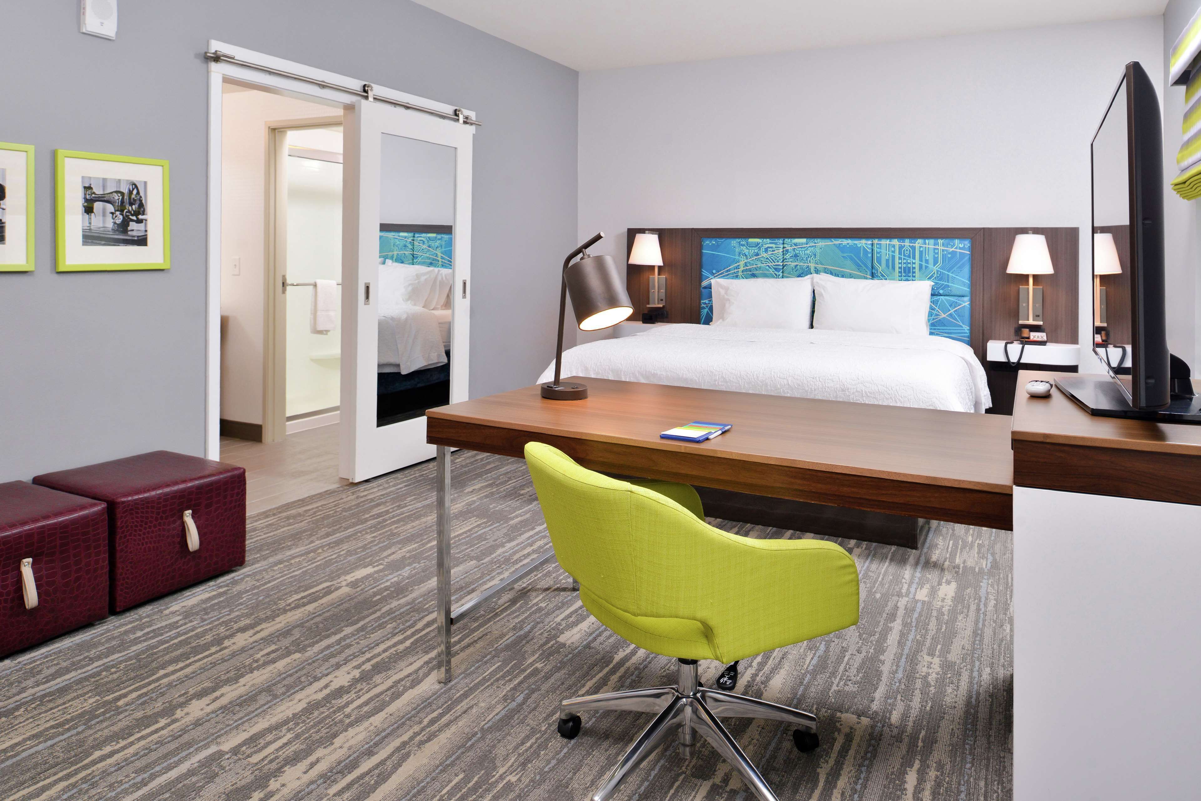 Habitación de hotel con cama, escritorio, silla y dos lámparas de noche. Se ve una puerta que da a otra habitación y una mesita con una lámpara.