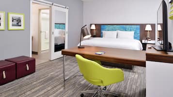 Habitación de hotel con cama, escritorio, silla y dos lámparas de noche. Se ve una puerta que da a otra habitación y una mesita con una lámpara.