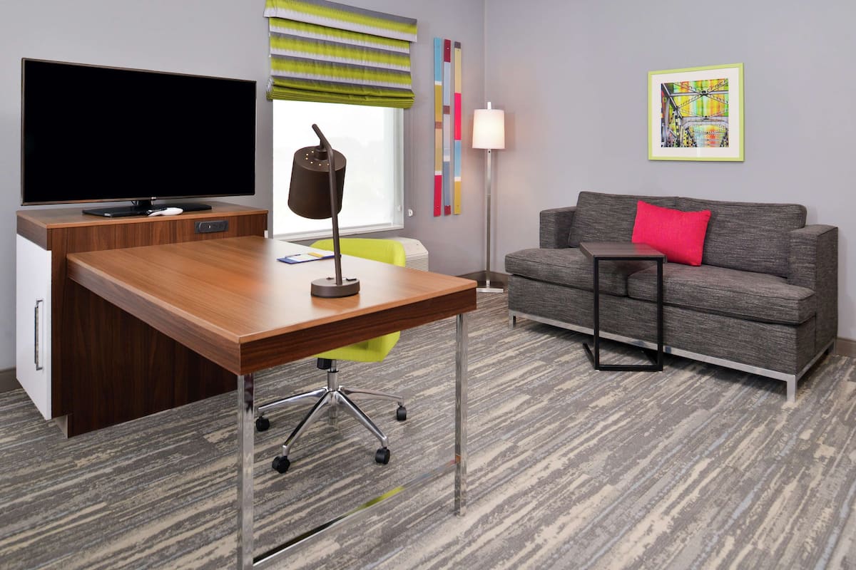 A modern room with a wooden desk, a flat-screen TV, a grey sofa with a red pillow, a standing lamp, and a colorful abstract painting on the wall.