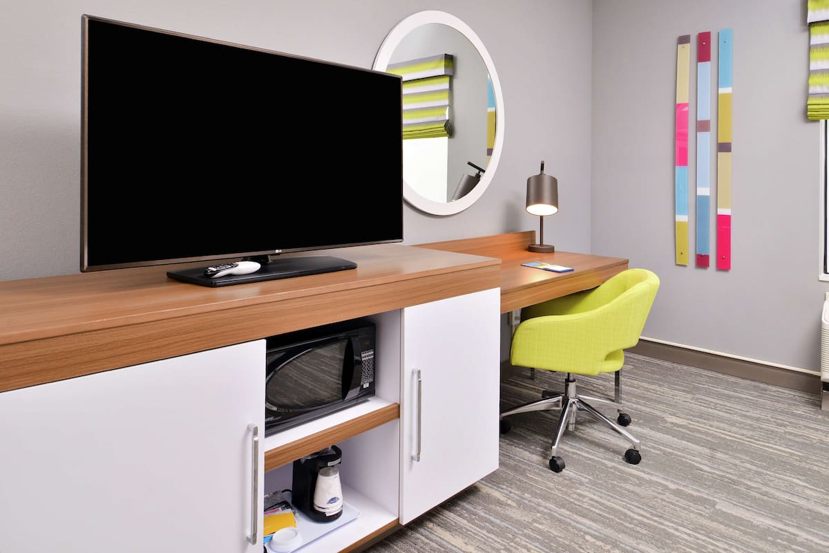 A modern hotel room with a wooden desk, a flat-screen TV, a microwave, a coffee maker, and a yellow chair.