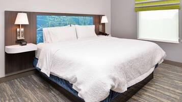 A neatly made bed with white linens, two bedside tables with lamps, a large window with blinds, and a wall-mounted artwork.