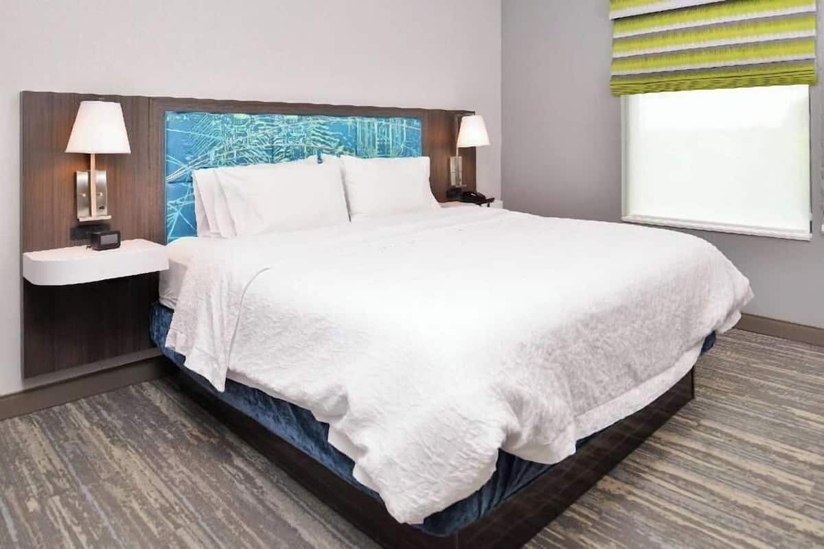 A hotel room with a large bed, two bedside tables with lamps, a headboard with a blue abstract design, and a window with a striped blind.