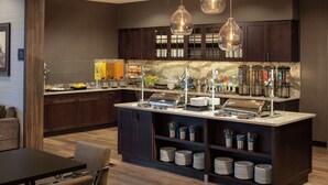A modern kitchen with dark wood cabinets, a central island with a glass top, and hanging pendant lights. - Homewood Suites by Hilton Needham Boston (Needham)