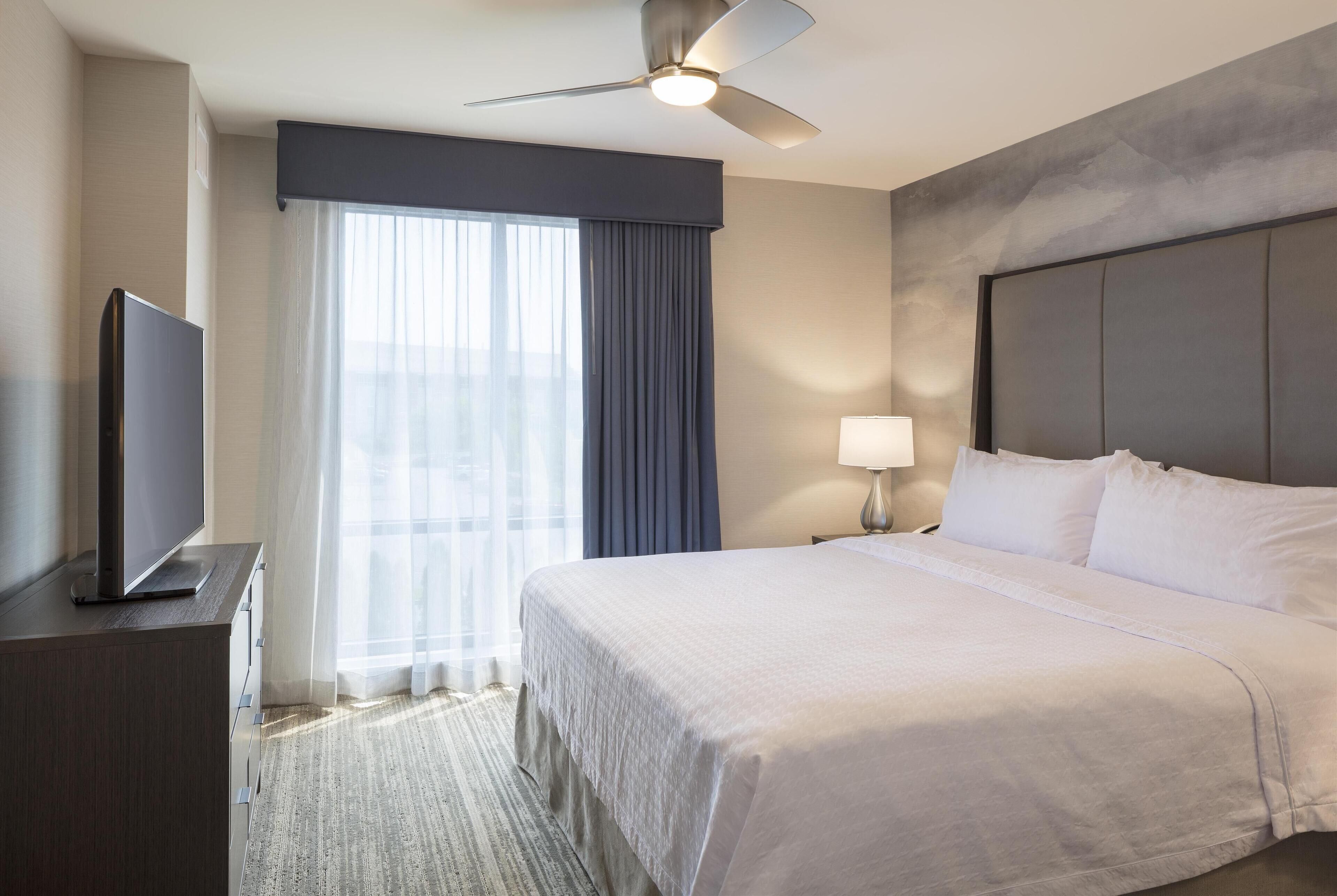 A hotel room with a bed, a TV on a stand, a ceiling fan, a window with curtains, and a nightstand with a lamp.
