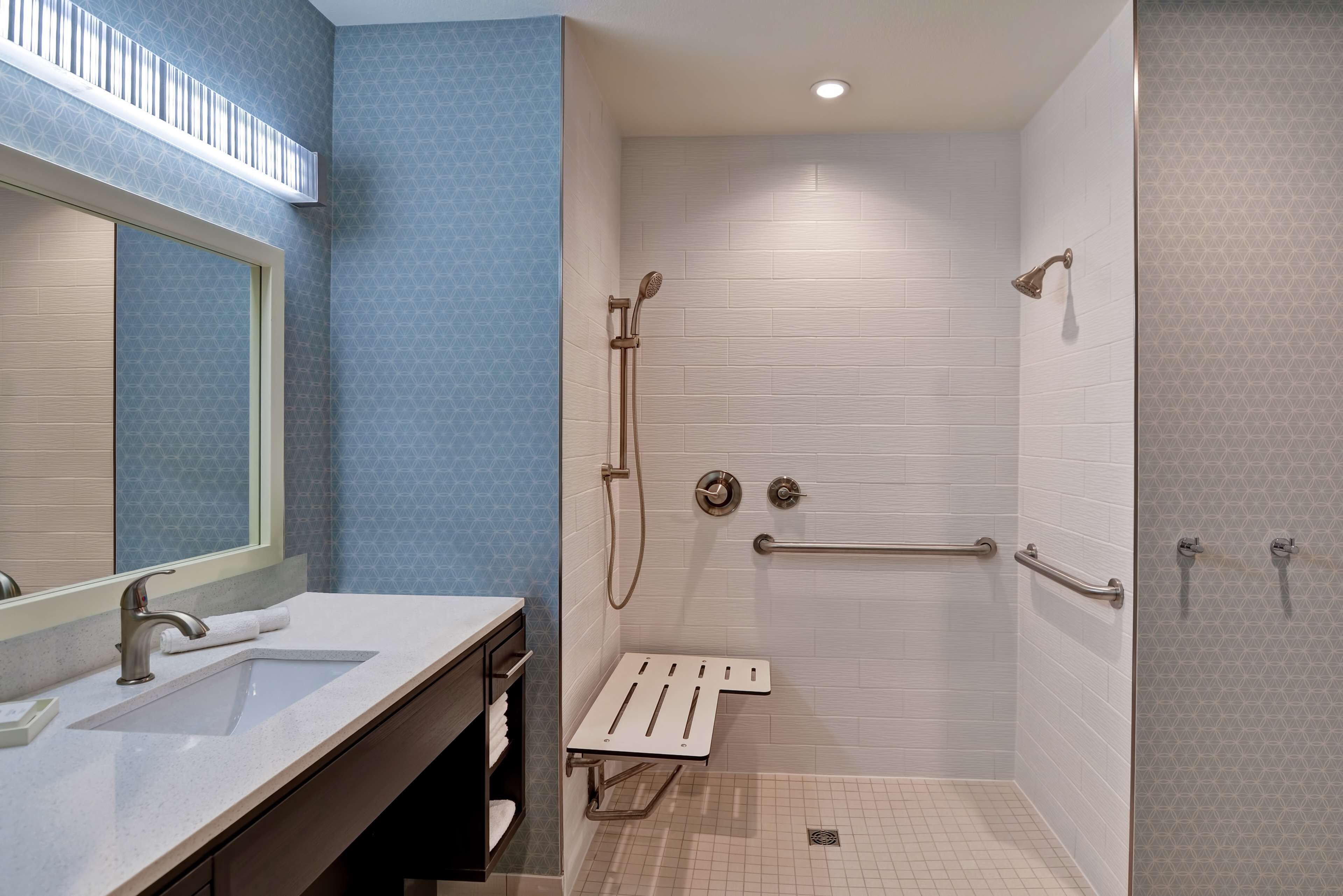 Une salle de bain moderne avec un grand miroir, deux vasques, une douche équipée de barres d’appui et un petit banc.