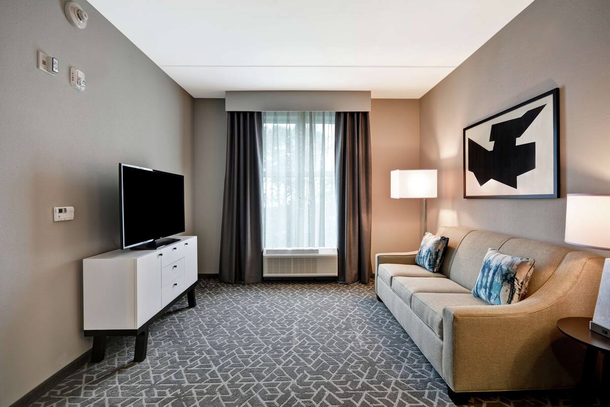 A hotel room with a flat-screen TV on a white cabinet, a beige sofa with patterned cushions, a large window with curtains, and a wall-mounted artwork.