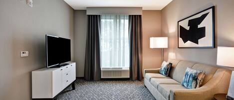 Hotelkamer met een flatscreen-televisie op een witte kast, een beige bank met decoratieve kussens, een groot raam met gordijnen en een kunstwerk aan de muur.
