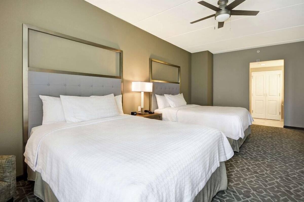 A hotel room with two beds, a ceiling fan, a nightstand with a lamp, and a door leading to another room.