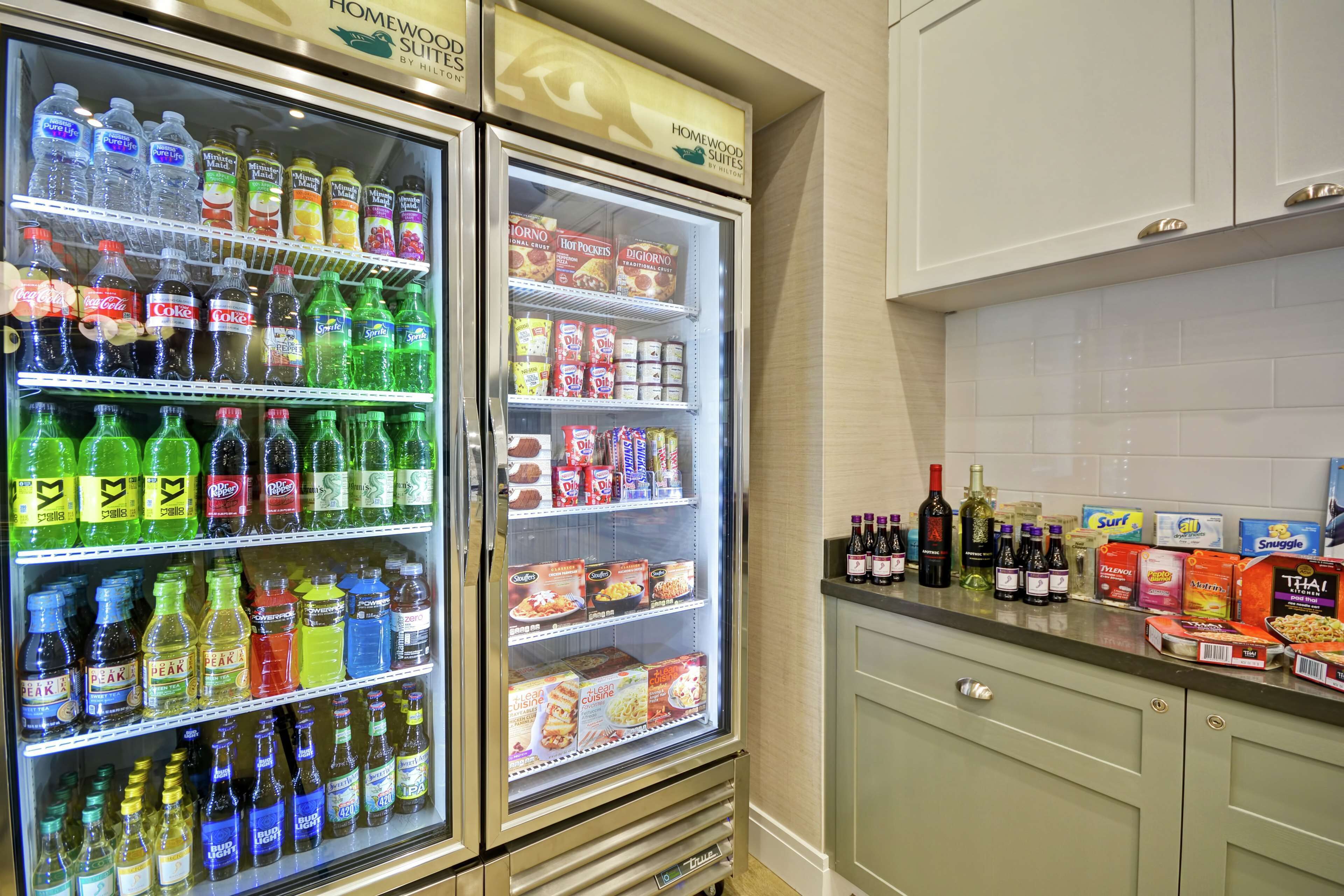 A kitchen with two refrigerators stocked with beverages and snacks, a countertop with various food items and bottles, and a cabinet with a handle.