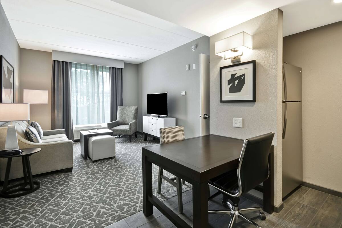 A modern hotel room with a dining area, a sofa, a TV, and a refrigerator.