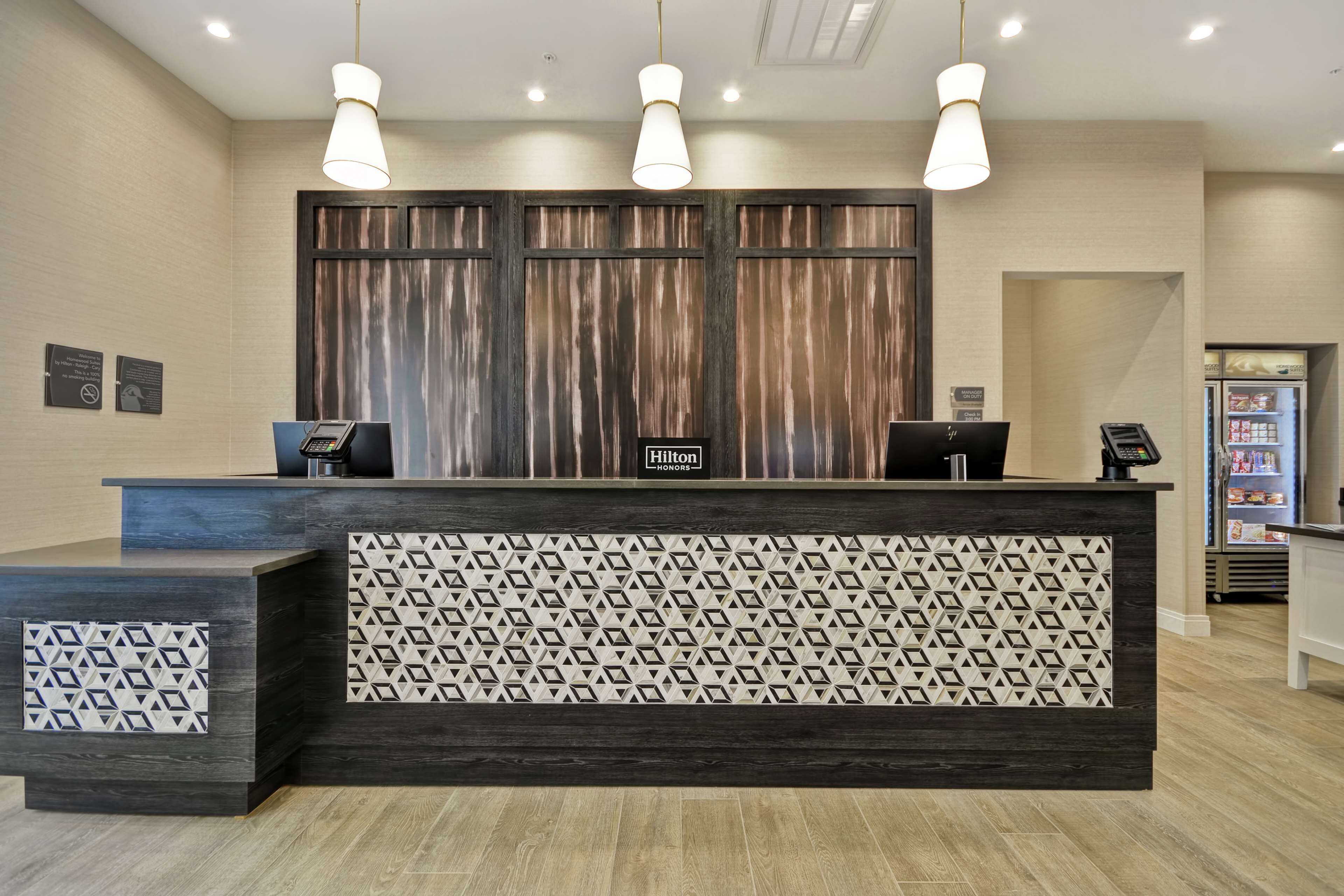 A hotel reception desk with a modern design, featuring a dark wood finish and patterned back panel. There are three pendant lights above the desk, and a minibar refrigerator is visible to the right.