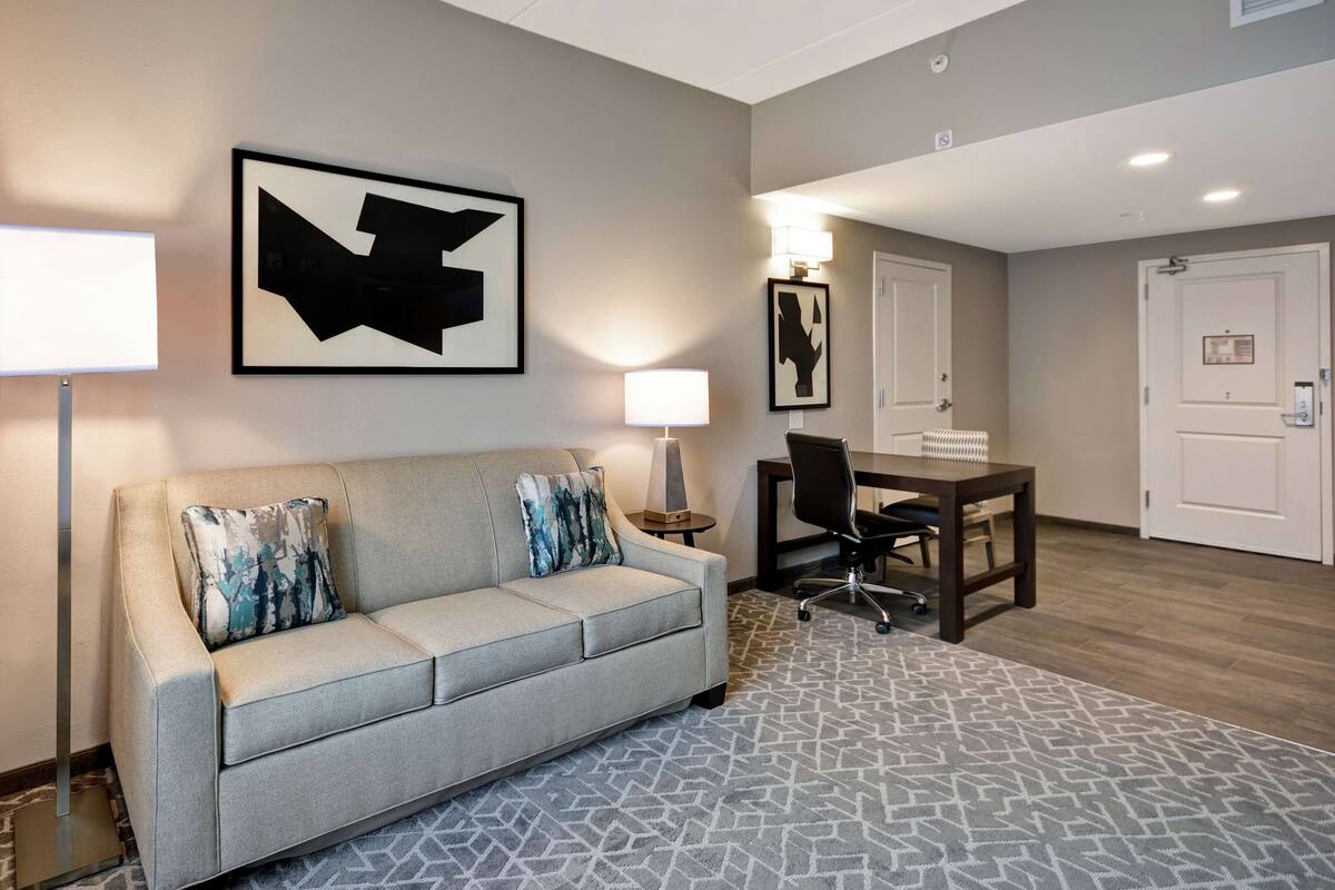 A modern hotel room with a grey sofa, a patterned rug, a desk with a chair, and framed artwork on the walls.