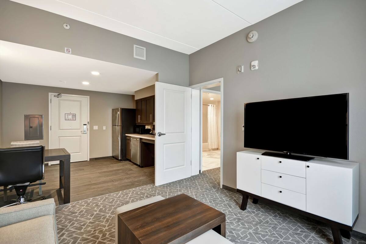 A modern living room with a flat-screen TV, a coffee table, and a sofa. There is a kitchen area with cabinets and a refrigerator.