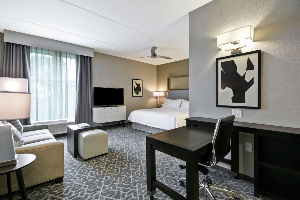 A hotel room with a bed, a desk, a chair, a TV, a lamp, and a painting on the wall.