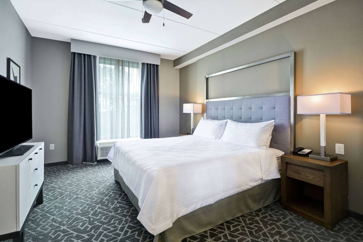 A hotel room with a bed, a TV, a bedside table with a lamp, and a window with curtains.