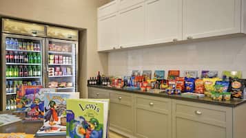 A kitchen area with a refrigerator stocked with beverages, a counter with snacks and books, and cabinets above.