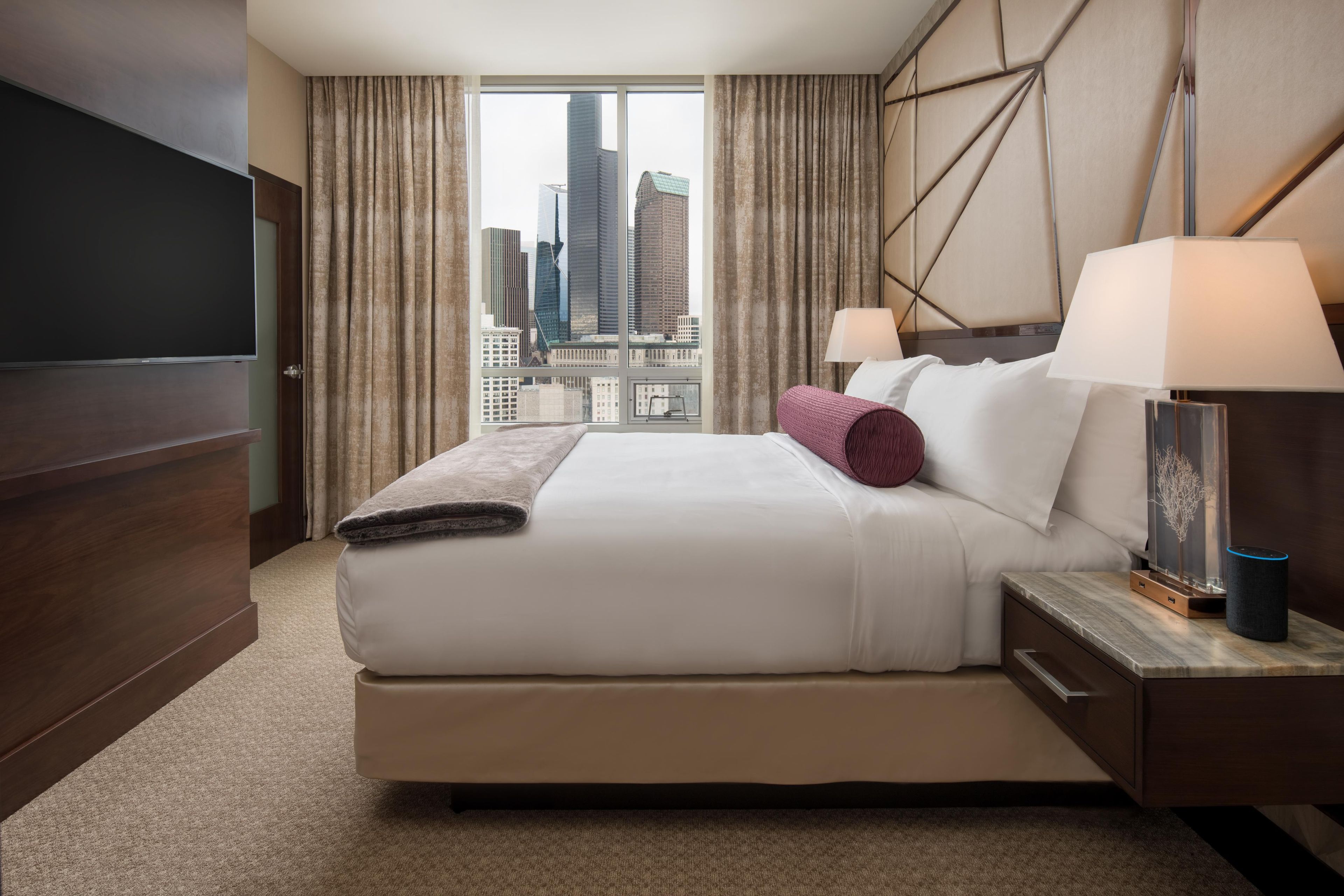 A hotel room with a large bed, a flat-screen TV, a nightstand with a lamp, and a view of the cityscape through the window.